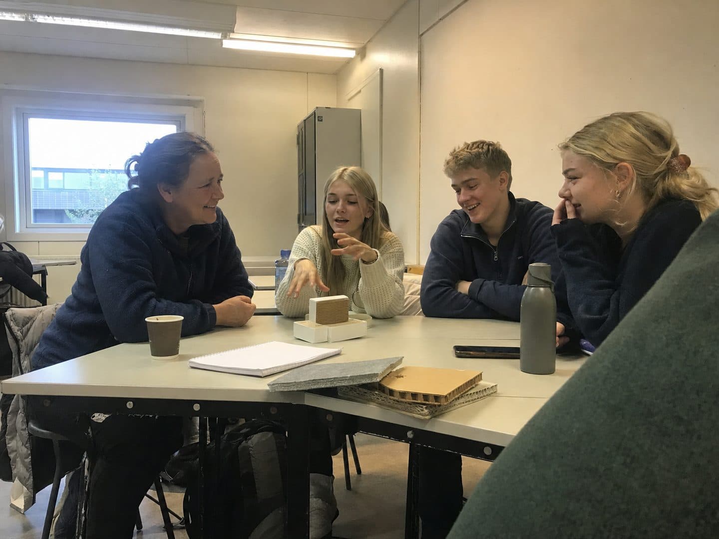En mentor sitter ved bordet med tre elever som forklarer ideen sin.