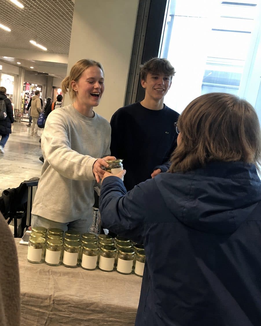 En elev rekker et glass til en kunde på messe og ler.
